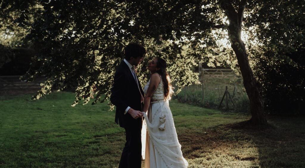 Bride and groom holding hands beneath a large tree in golden evening sunlight at Hall Farm, Norfolk, with a wooden field gate behind them