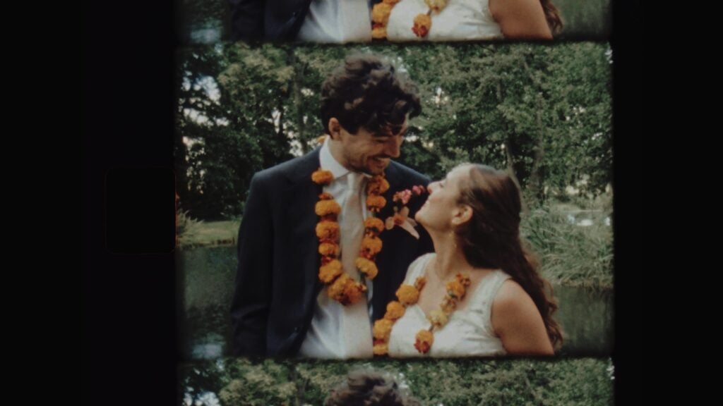 Super 8 film still of the bride and groom in marigold garlands laughing together beside a pond surrounded by trees