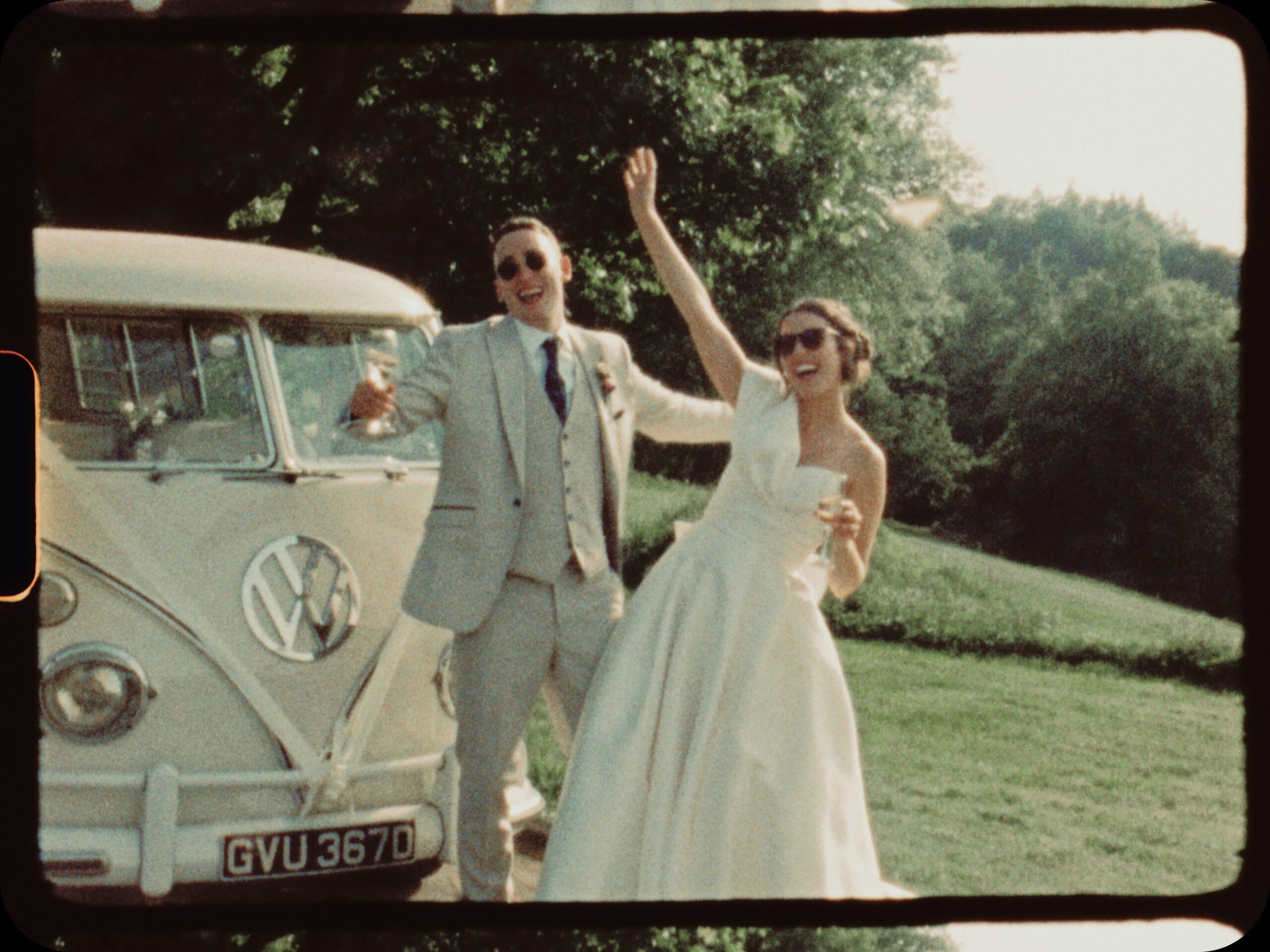 Super 8 film still of the bride and groom in sunglasses waving and laughing beside a cream vintage VW campervan on a sunny lawn