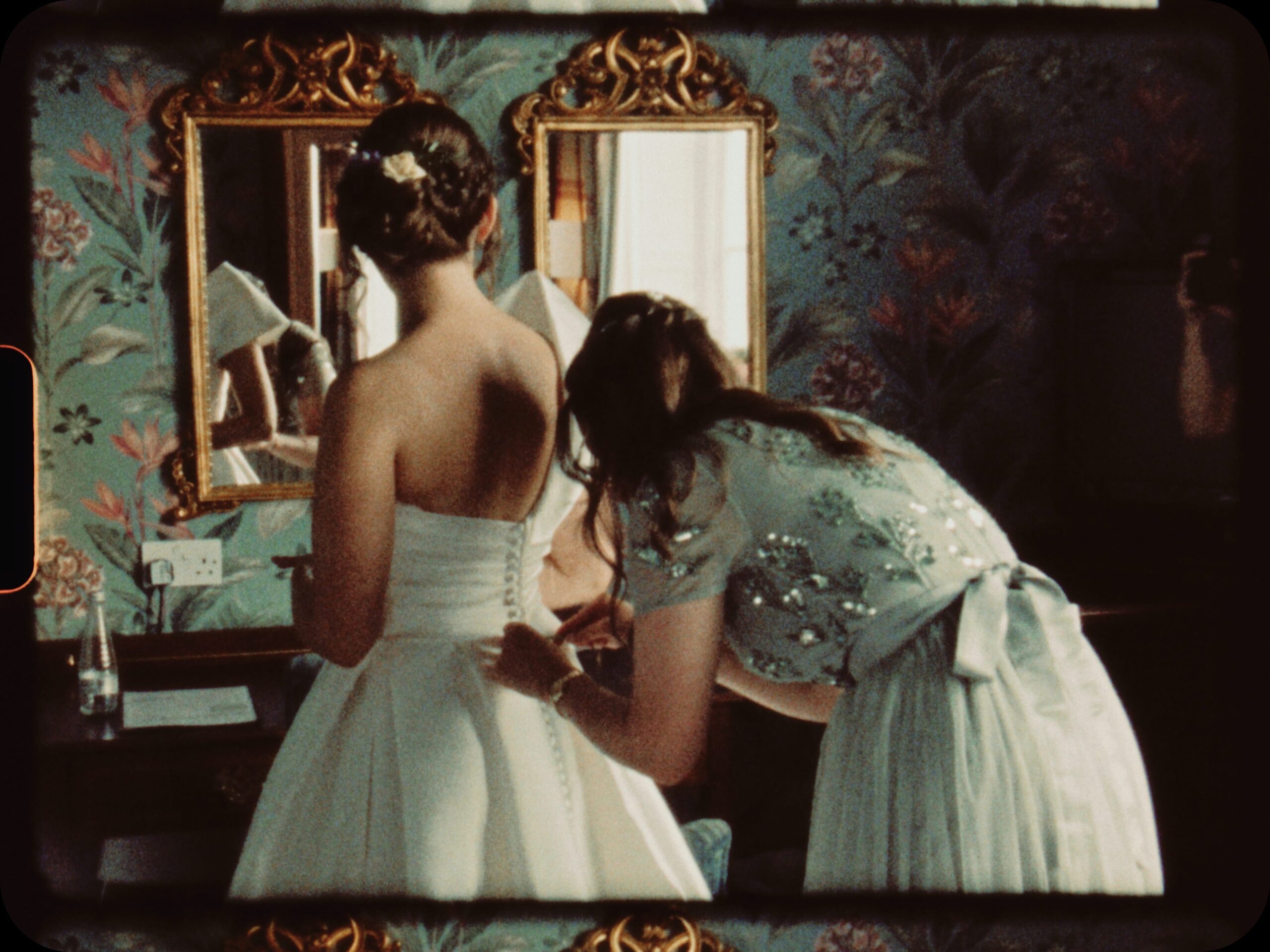 Super 8 film still of a bridesmaid buttoning the back of the bride’s dress in front of gold-framed mirrors and bold floral wallpaper