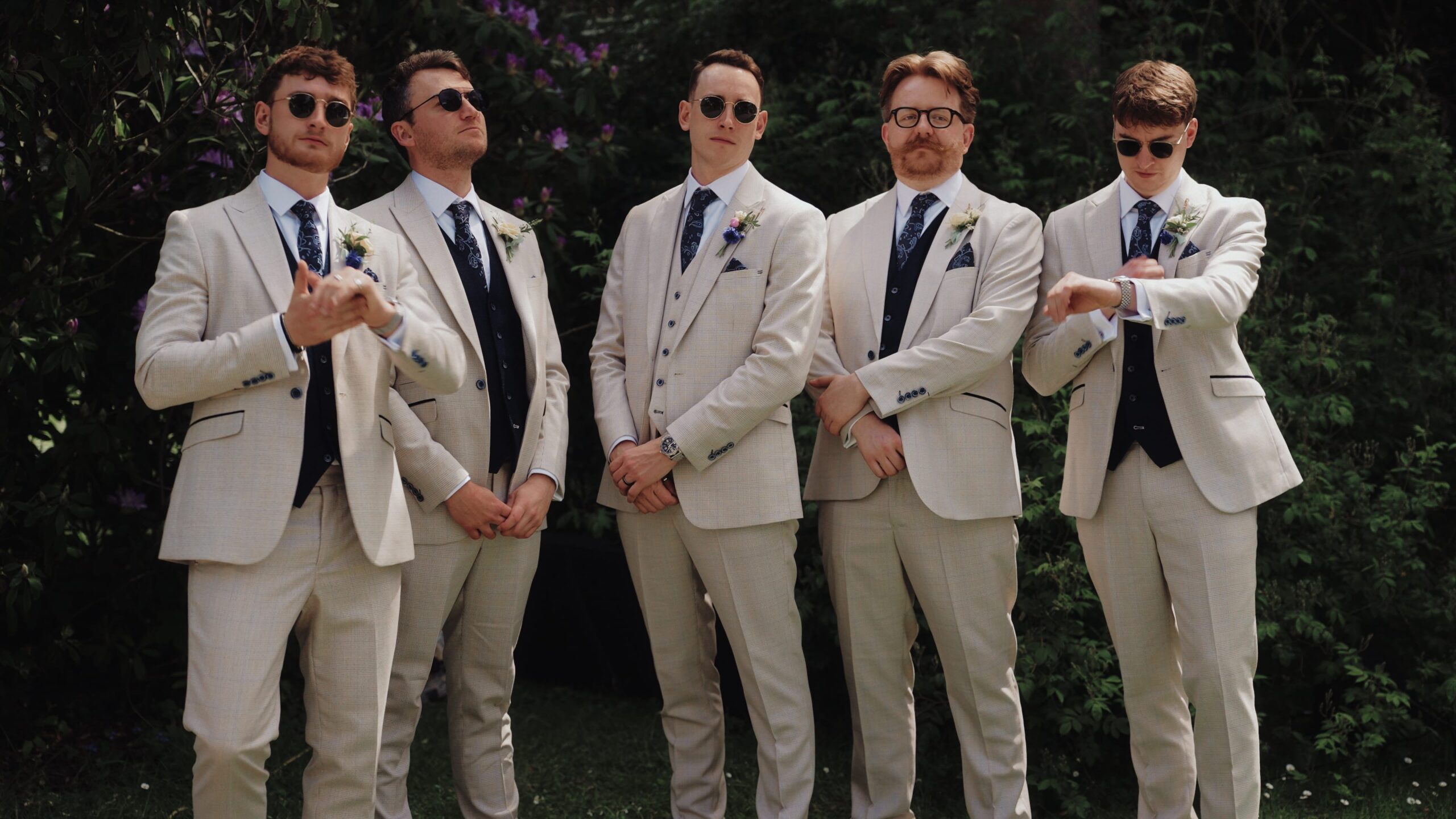Groom and four groomsmen in matching cream suits and round sunglasses striking playful poses in front of rhododendron bushes