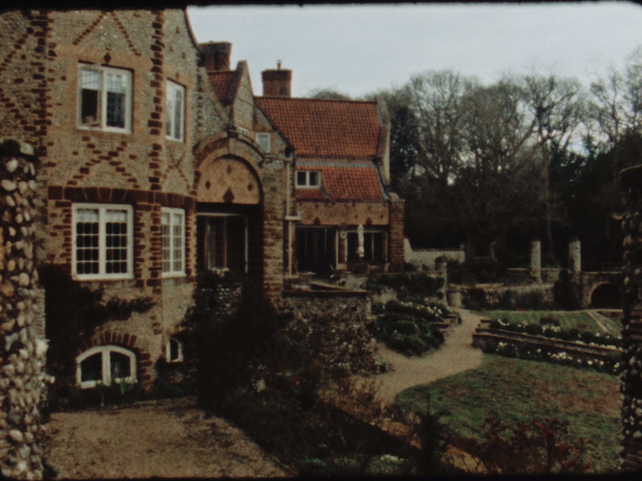 Super 8 film still of Voewood’s flint and brick Arts and Crafts house in Norfolk, looking across to a winding gravel path and sunken garden