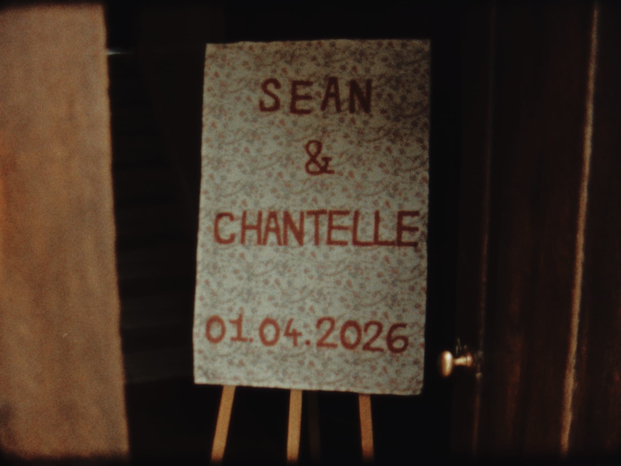 Super 8 film still of a handmade floral wedding sign with the couple’s names and date, displayed on an easel inside Voewood
