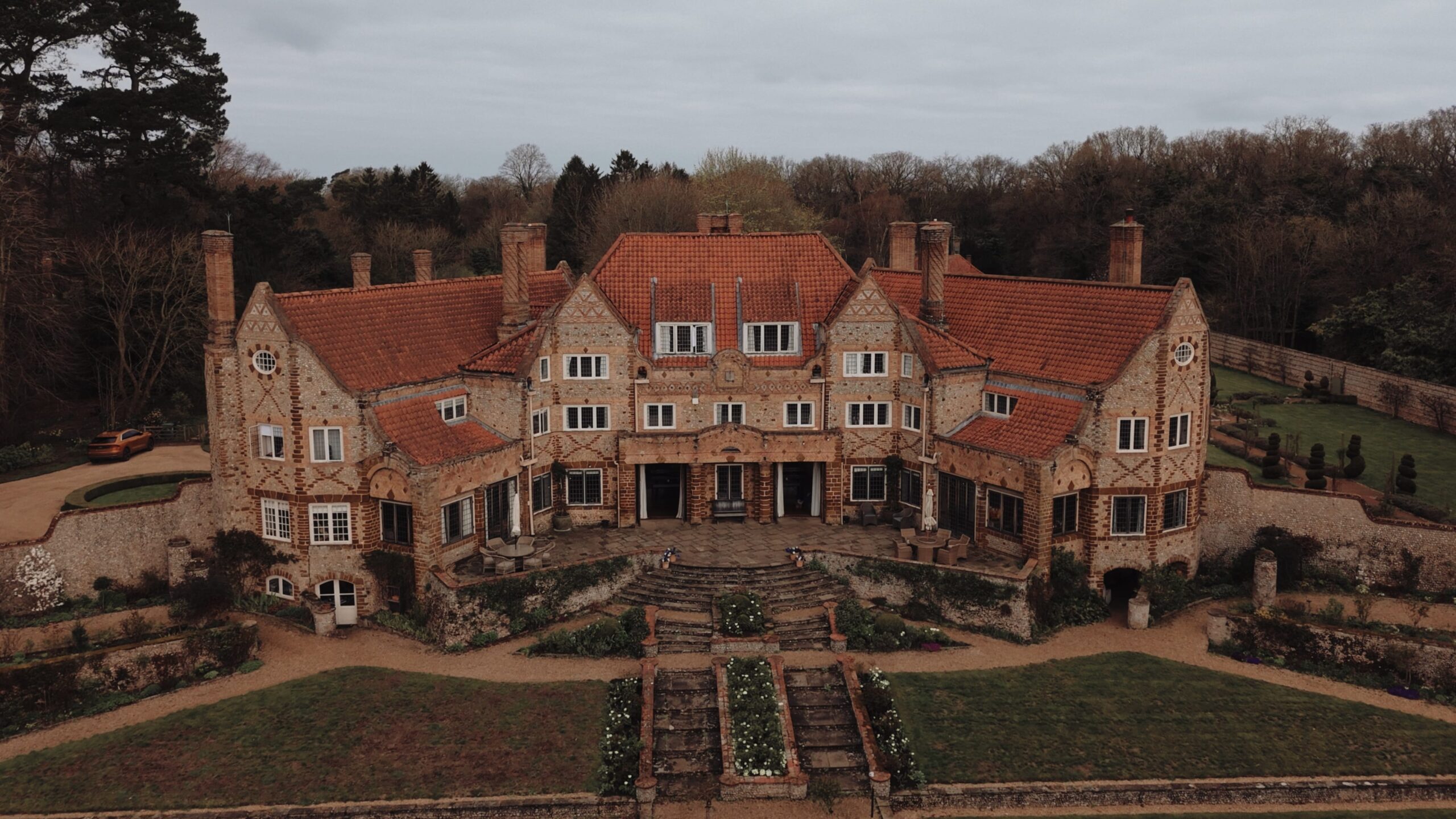 Aerial view of Voewood in Norfolk, showing the flint and brick Arts and Crafts house with terracotta roofs, curved stone steps down to the terraced lawn and woodland behind